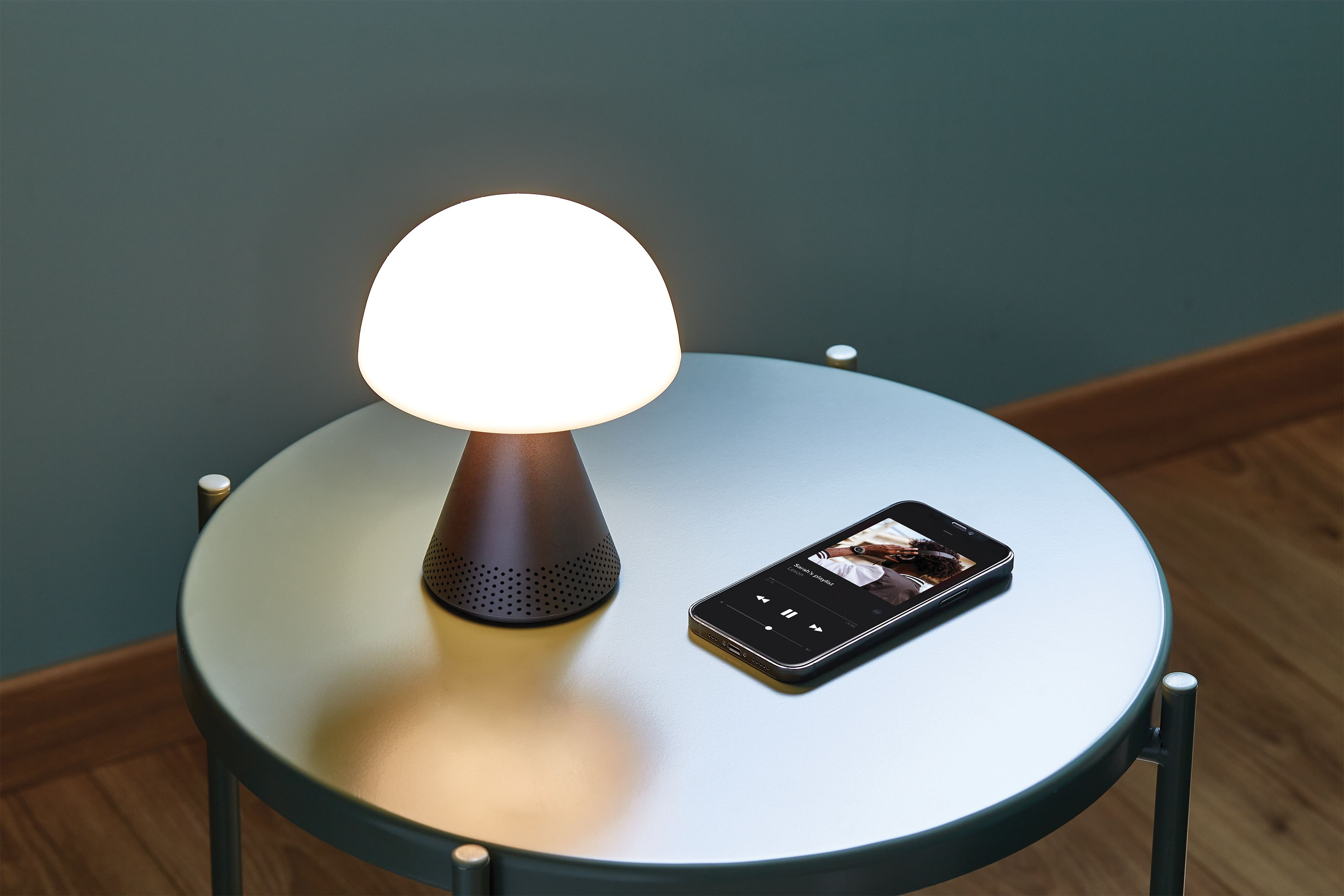 Lampe de table et téléphone sur une table. La lampe est blanche et marron. Le téléphone affiche une application musicale.