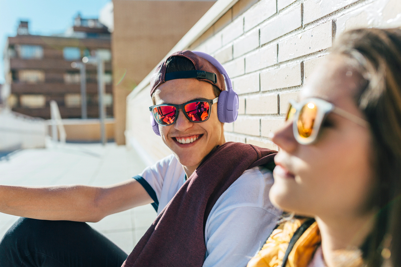 Eine Person mit Kopfhörern und Sonnenbrille lächelt in der Sonne, eine andere Person im Hintergrund.
