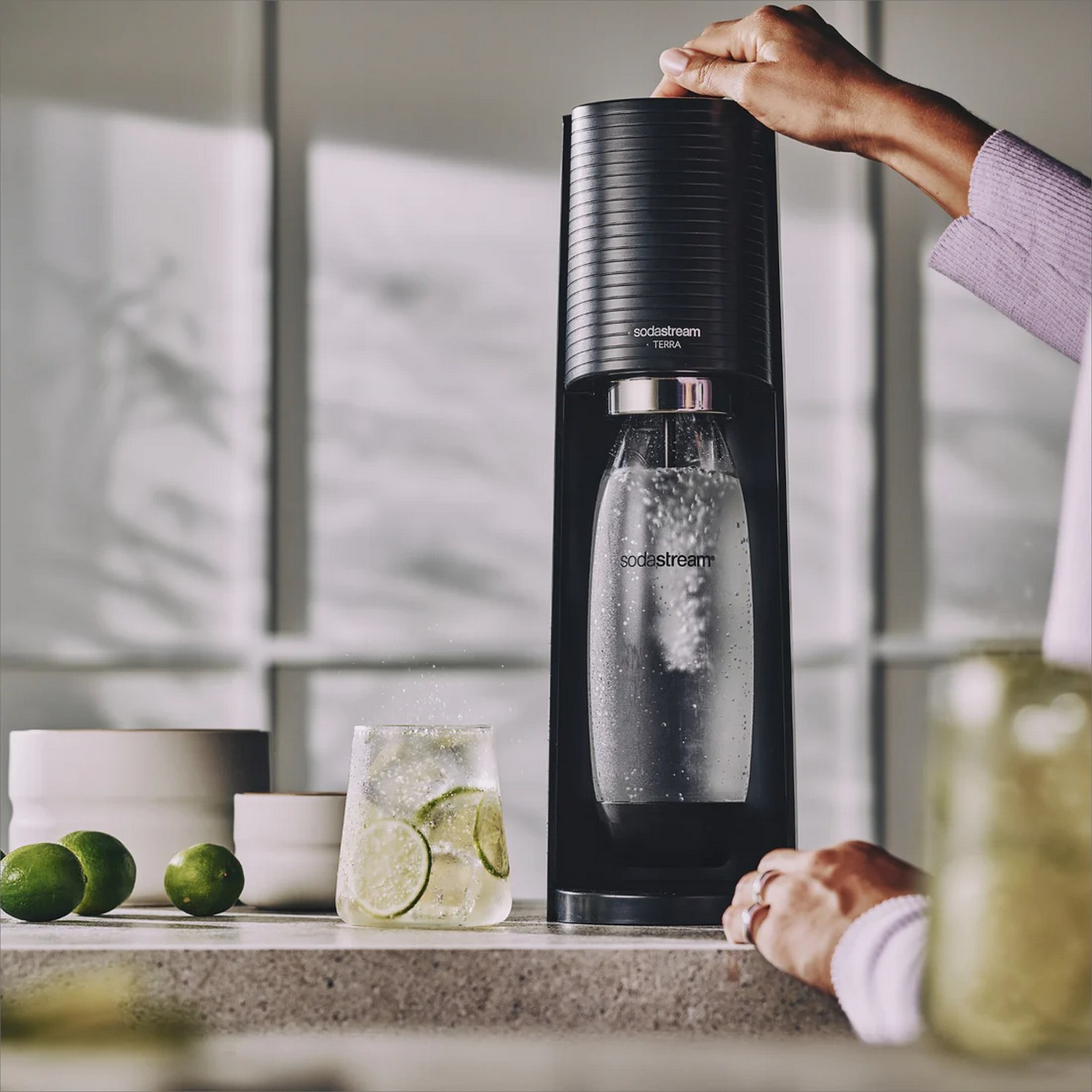 Une machine SodaStream noire avec un verre de boisson au citron vert et des citrons verts sur un comptoir.