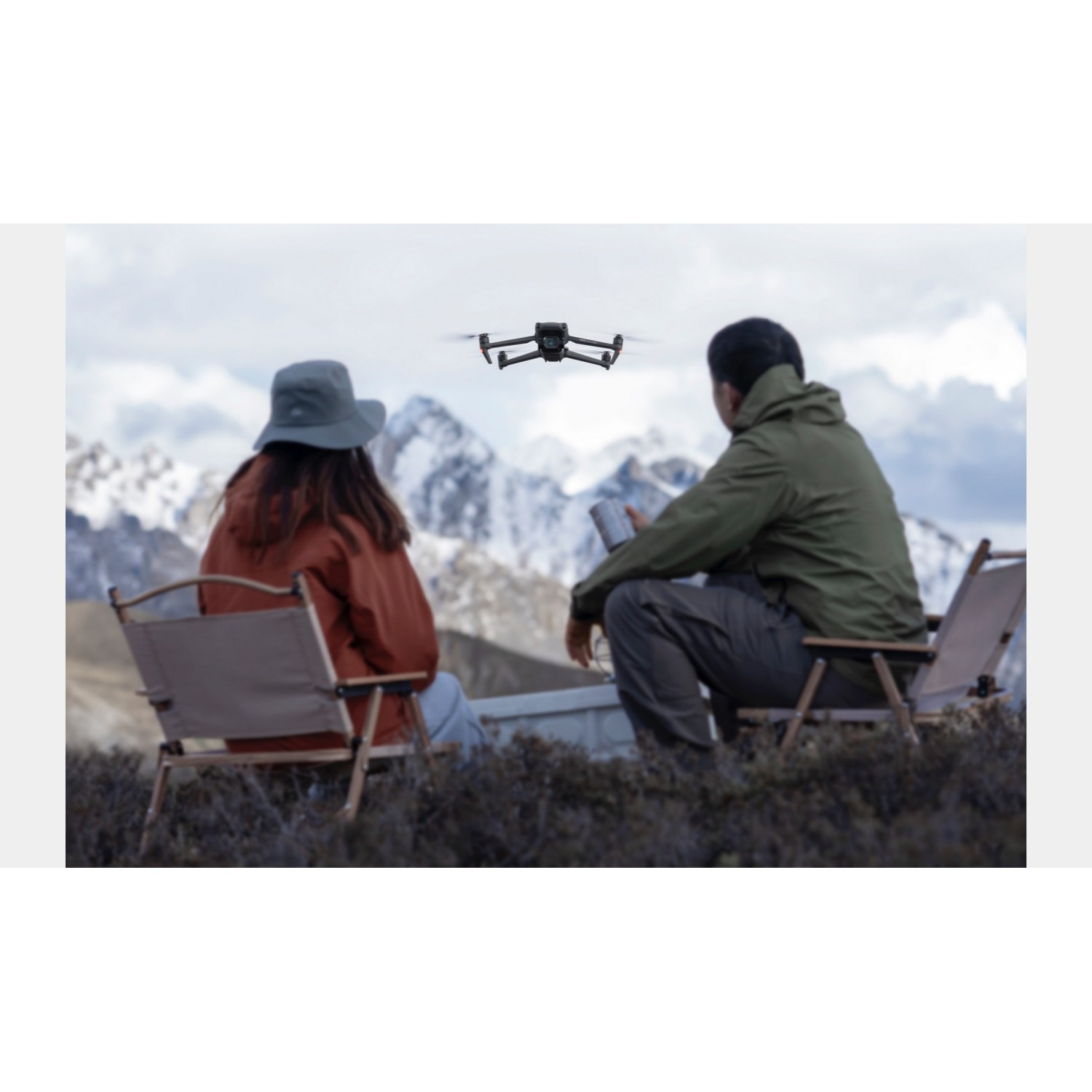 Een drone die over twee mensen vliegt die in stoelen zitten, besneeuwde bergen op de achtergrond.