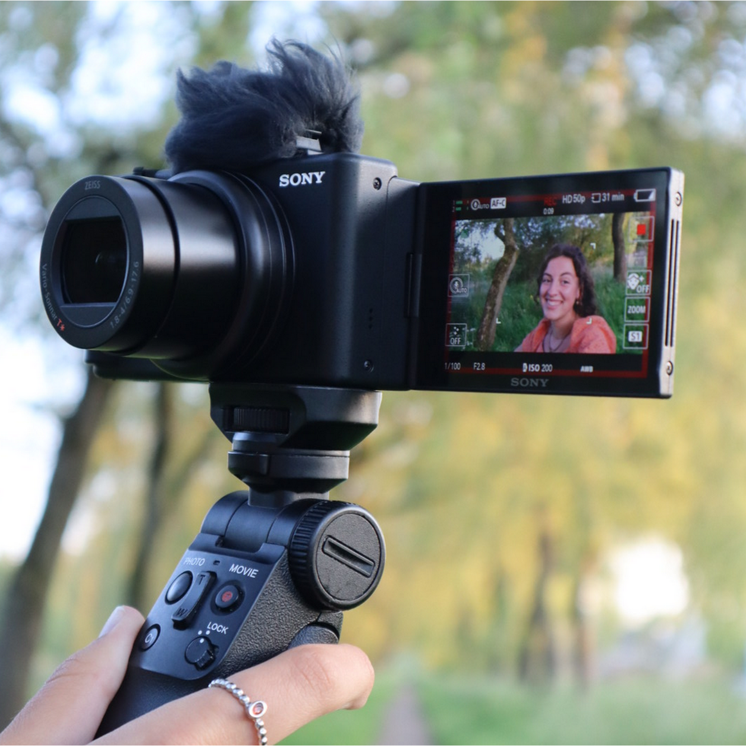 Woman filming herself with a camera on a tripod. Trees and a sunny day are in the background.