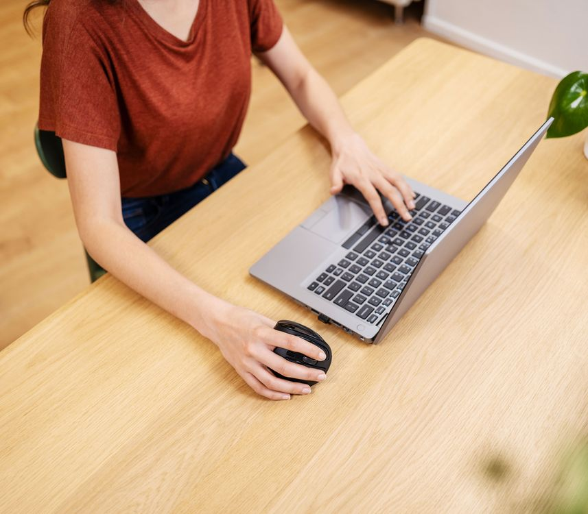Nő laptopot használ, egérrel a fa asztalon, növénnyel, felülnézet, a kezekre fókuszálva.