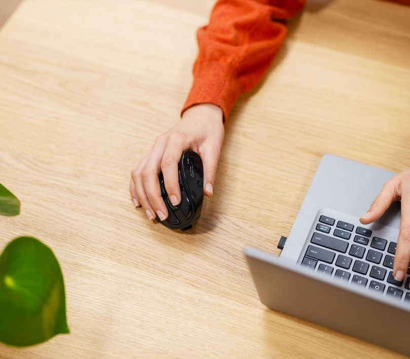 Egy személy keze egy fekete egeret használ egy laptop mellett egy fa asztalon egy növénnyel.