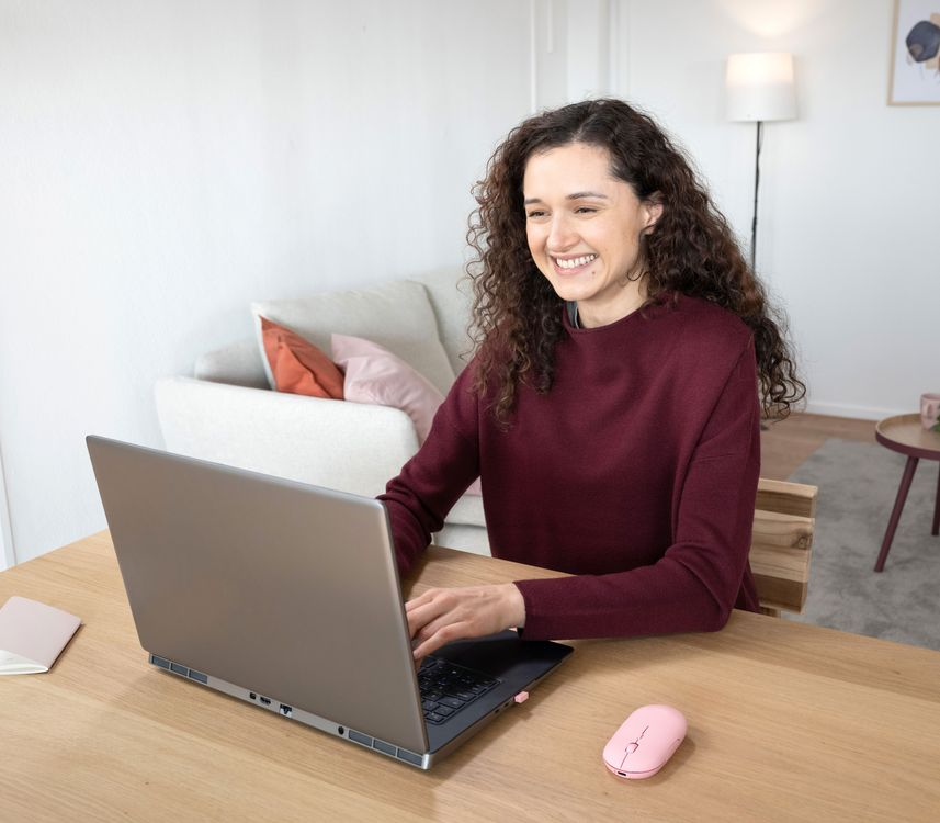 Nő mosolyogva dolgozik egy laptopon egy faasztalnál. Egy rózsaszín egér van a laptop mellett.