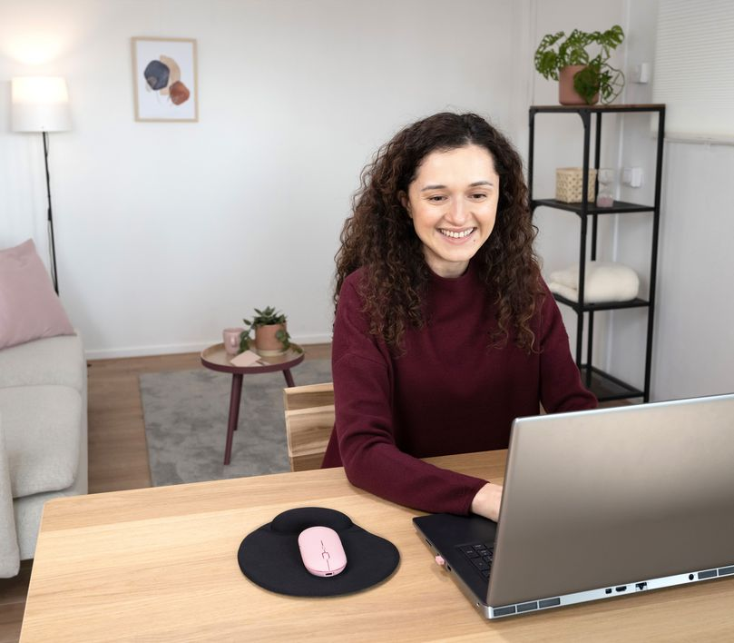 Egy nő mosolyog, miközben egy laptopon dolgozik egy fa asztalnál. Egy egér és egy egérpad van előtte.