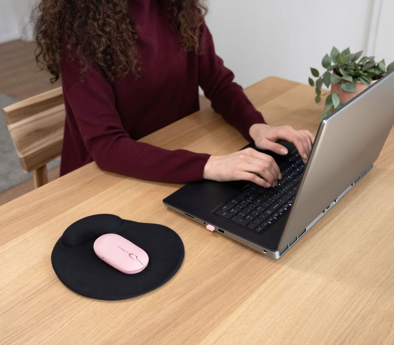 Nő laptopot használ. Rózsaszín egér és fekete egérpad. Fa asztal.