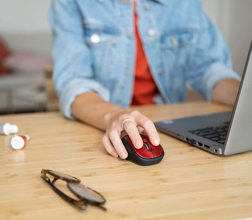 Személy egy piros számítógépes egeret használ, laptop és szemüveg az asztalon.