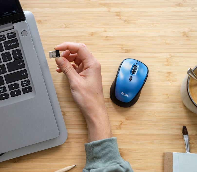 Egy kéz egy USB-vevőt tart egy kék egér és egy laptop mellett egy fa asztalon.