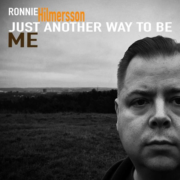 Album borító egy férfi arcával, a címe: 'Just Another Way To Be Me'. A háttérben mező, fák és egy város felhős ég alatt látható.