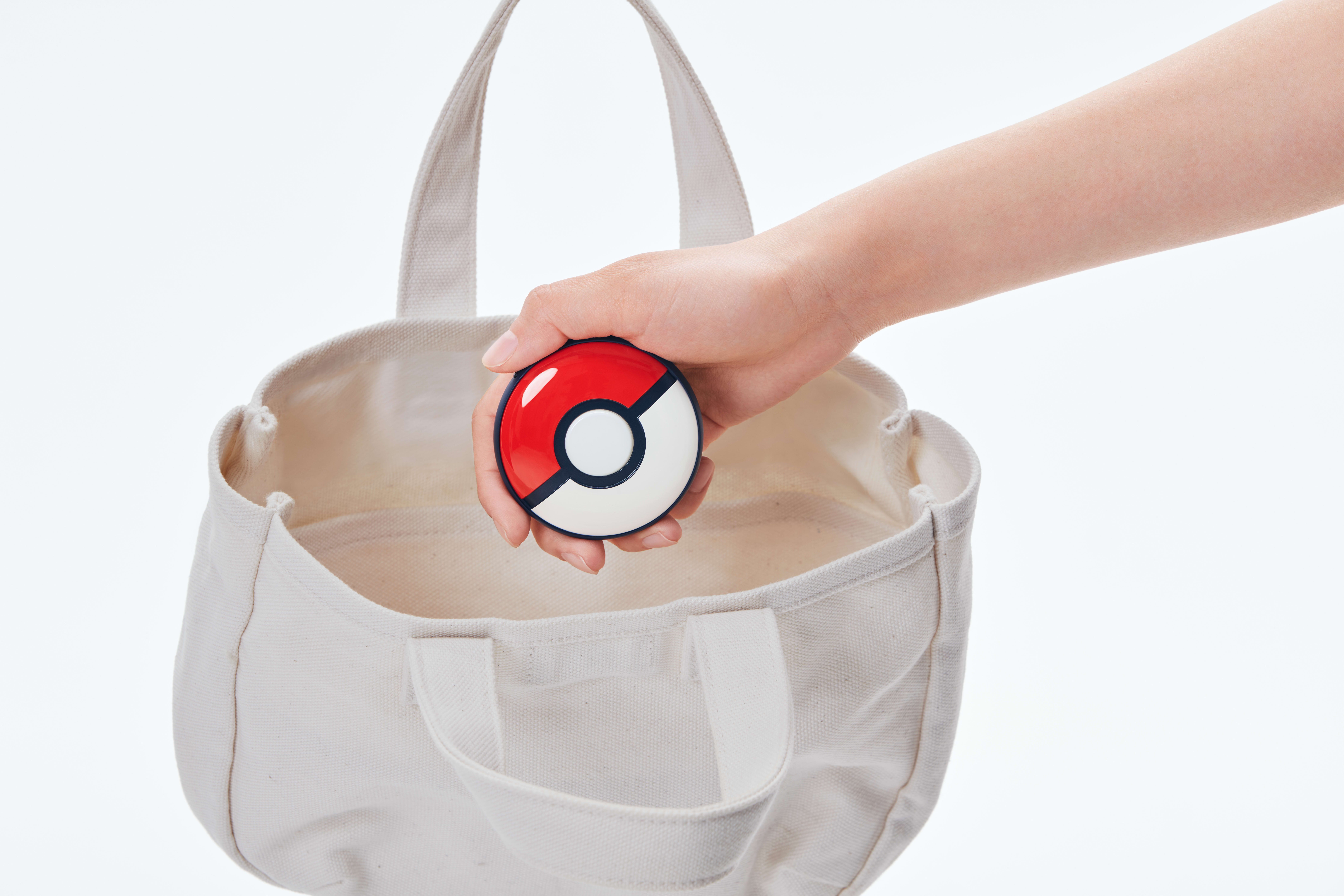 Eine Hand, die einen rot-weißen Pokéball in eine weiße Leinwandtasche legt. Die Tasche liegt auf weißem Grund.