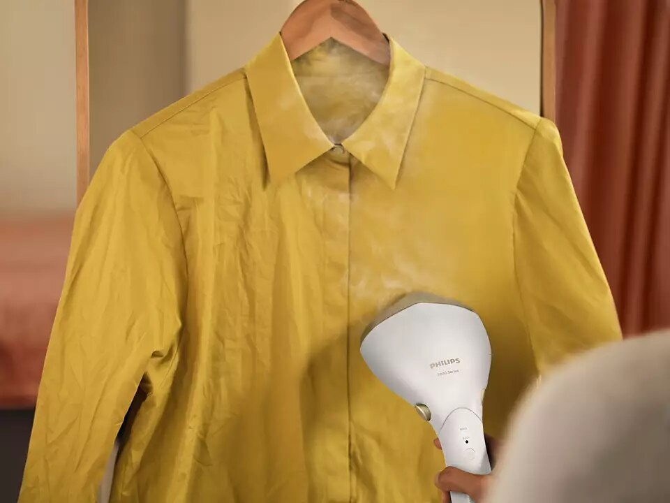 Osoba prasująca żółtą koszulę wiszącą na drewnianym wieszaku parownicą Philips.