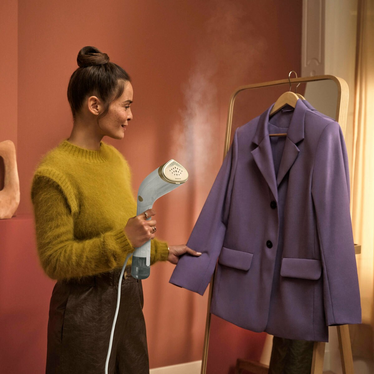 Kobieta paruje fioletową marynarkę na wieszaku. Ma na sobie żółty sweter i brązowe spodnie.