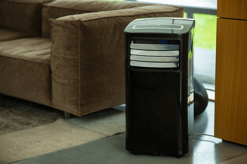 Zwarte draagbare airconditioner naast een bank. Het apparaat heeft zilveren en zwarte roosters en een bedieningspaneel.