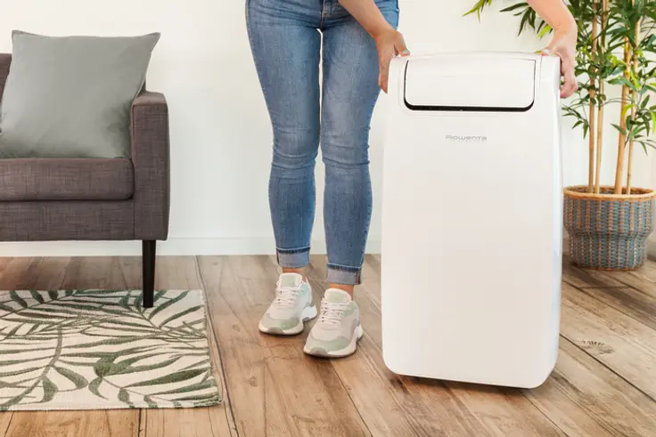 Een vrouw die een witte airconditioner vasthoudt. De airconditioner staat op een houten vloer.
