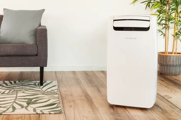 Witte airconditioner naast een grijze bank en een plant op een houten vloer, witte muur.