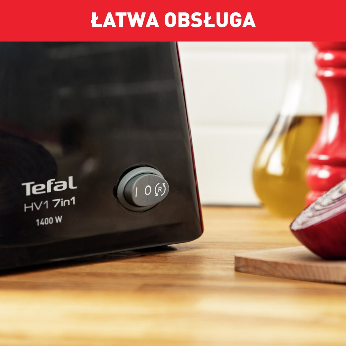 Czarny robot kuchenny z przyciskiem, cebulą i butelką oleju na drewnianej powierzchni.