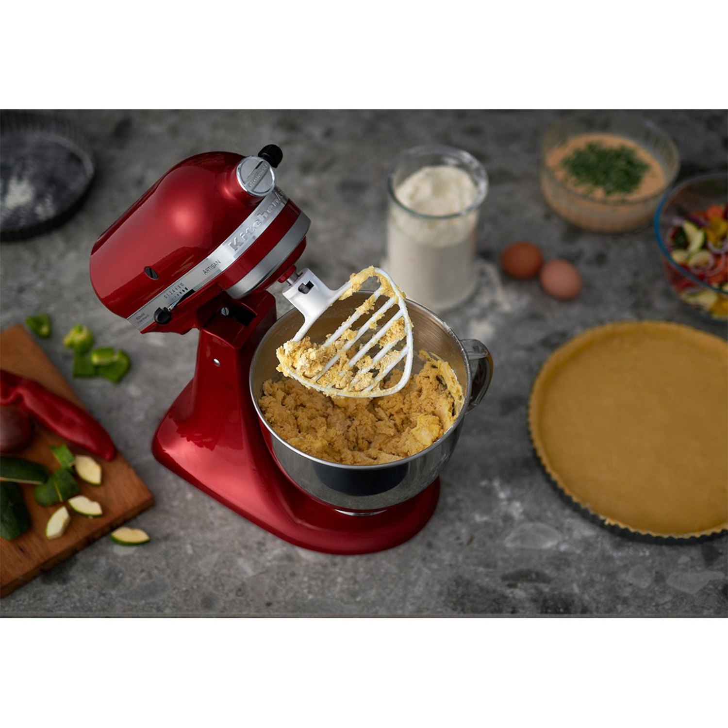 Impastatrice KitchenAid rossa con ciotola di impasto. Ingredienti e pasta per torta sullo sfondo.