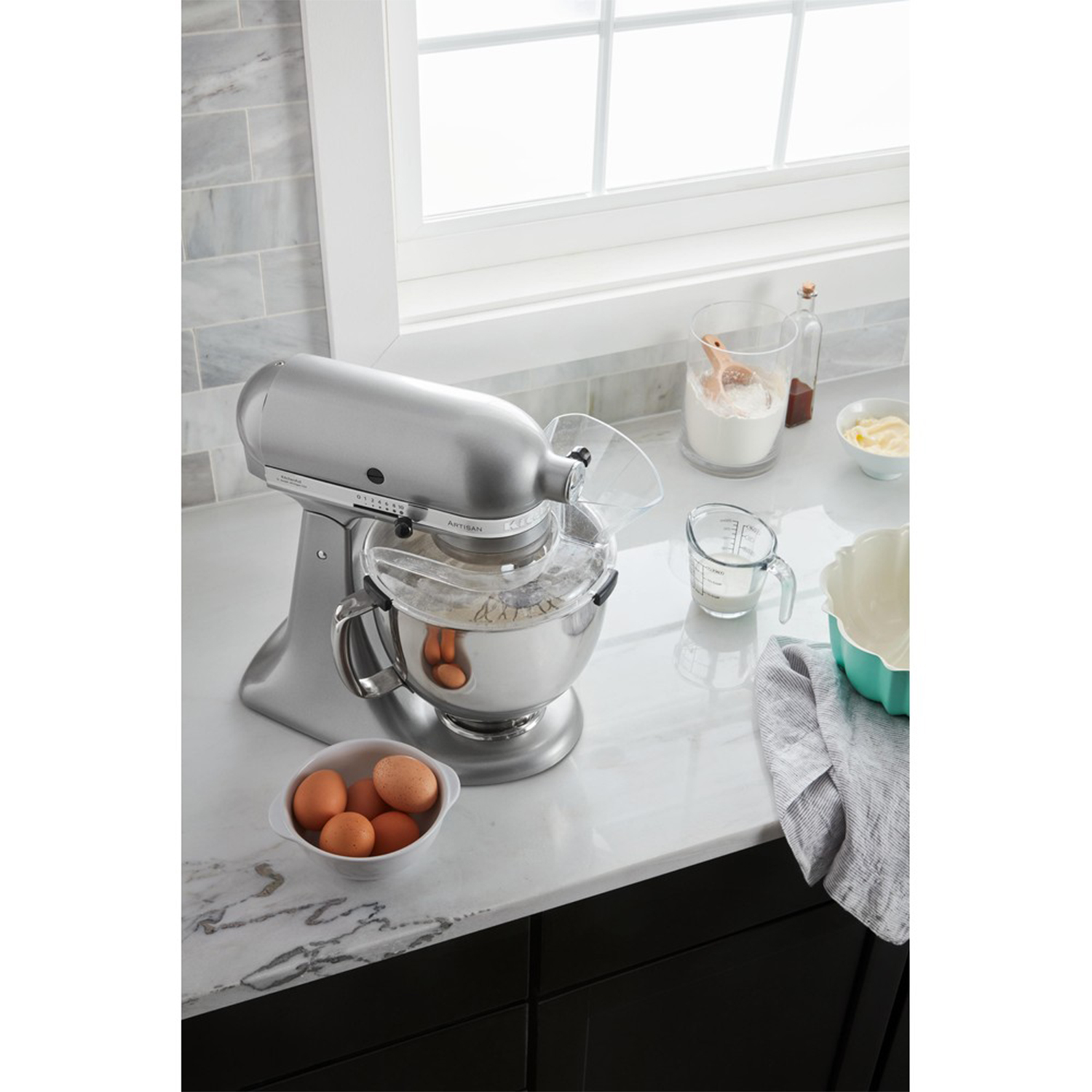 Un mixer KitchenAid argentato con ingredienti su un piano di lavoro in marmo vicino a una finestra.