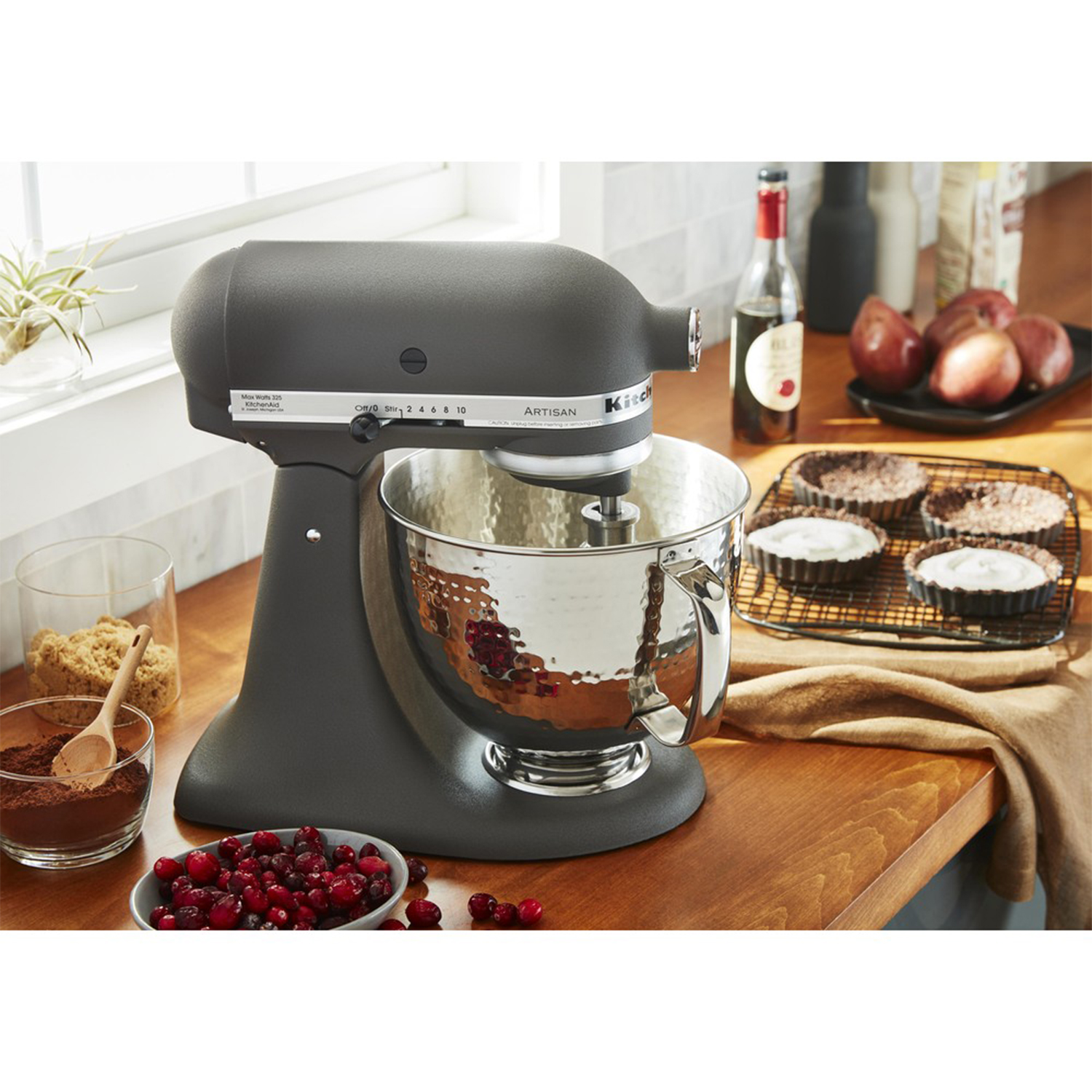 Un frullatore a immersione KitchenAid nero con una ciotola in acciaio inossidabile, accanto a ingredienti e crostate al forno.