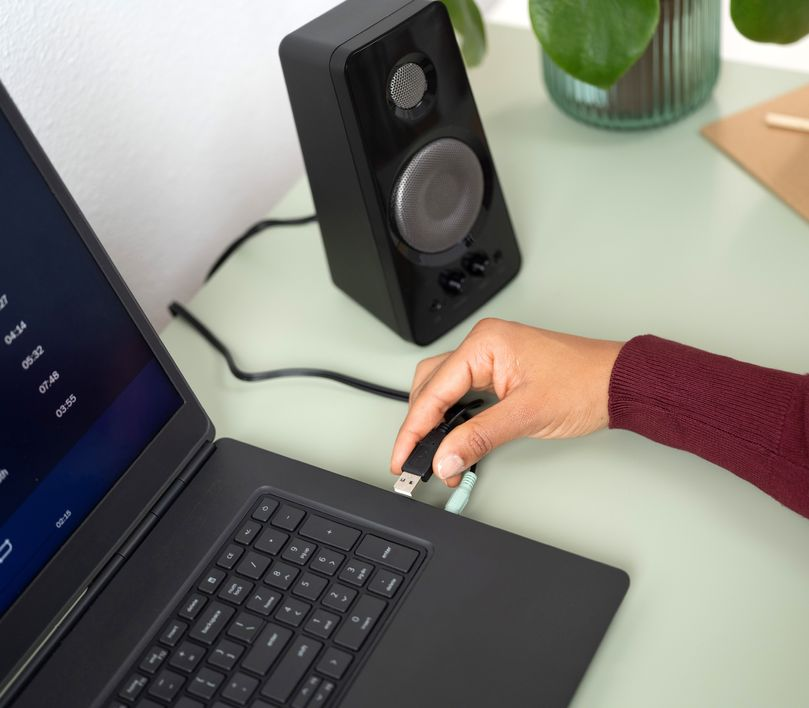 Fekete hangszóró egy laptop mellett, egy kézzel, amely egy USB-kábelt csatlakoztat.