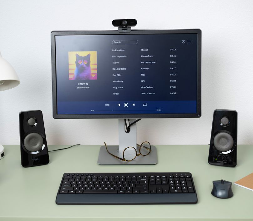 Egy asztali beállítás monitorral, hangszórókkal, billentyűzettel, egérrel és webkamerával.