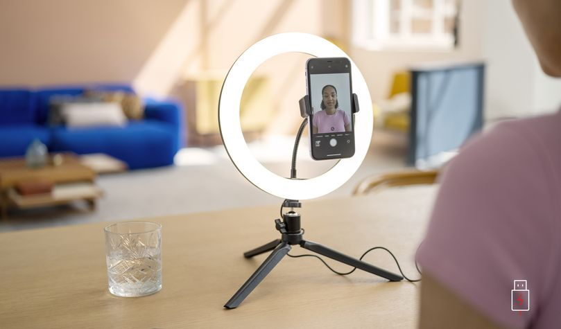 Egy személy telefonnal filmez egy gyűrűs lámpában. Egy pohár víz van az asztalon.