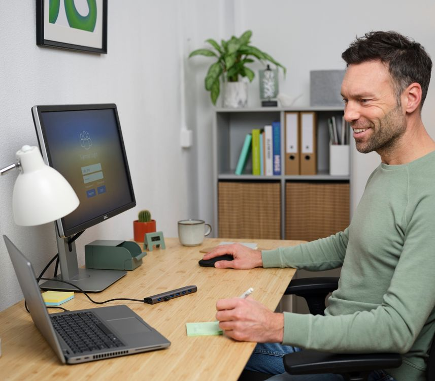 Férfi dolgozik egy asztalnál laptop, monitor és toll segítségével. Mosolyog, zöld pulóverben.