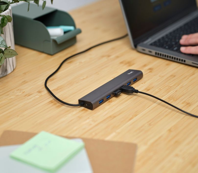 Asztal laptopral és csatlakoztatott USB hubbal. Van egy zöld post-it és egy növény a háttérben.