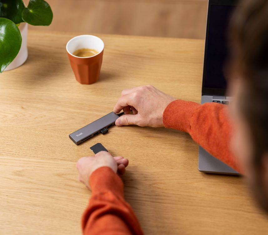 Személy kezei USB-elosztóval és SD-kártyával egy faasztalon, laptop és kávé közelében.