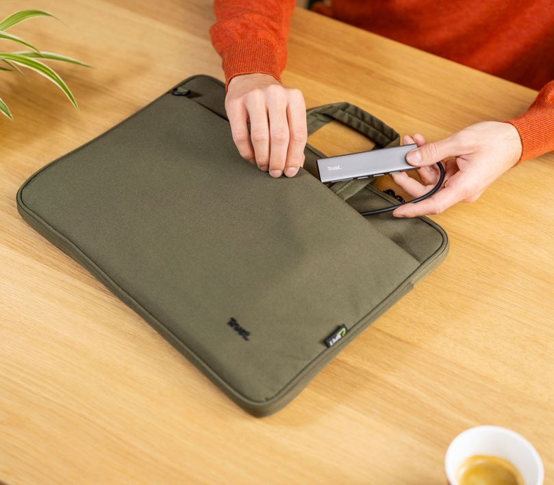 Egy személy kezei egy szürke hub-ot helyeznek egy zöld laptop tokba a faasztalon.