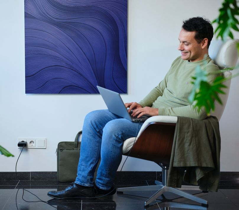 Férfi egy széken ül, és laptopon dolgozik. Művészet van a mögötte lévő falon.