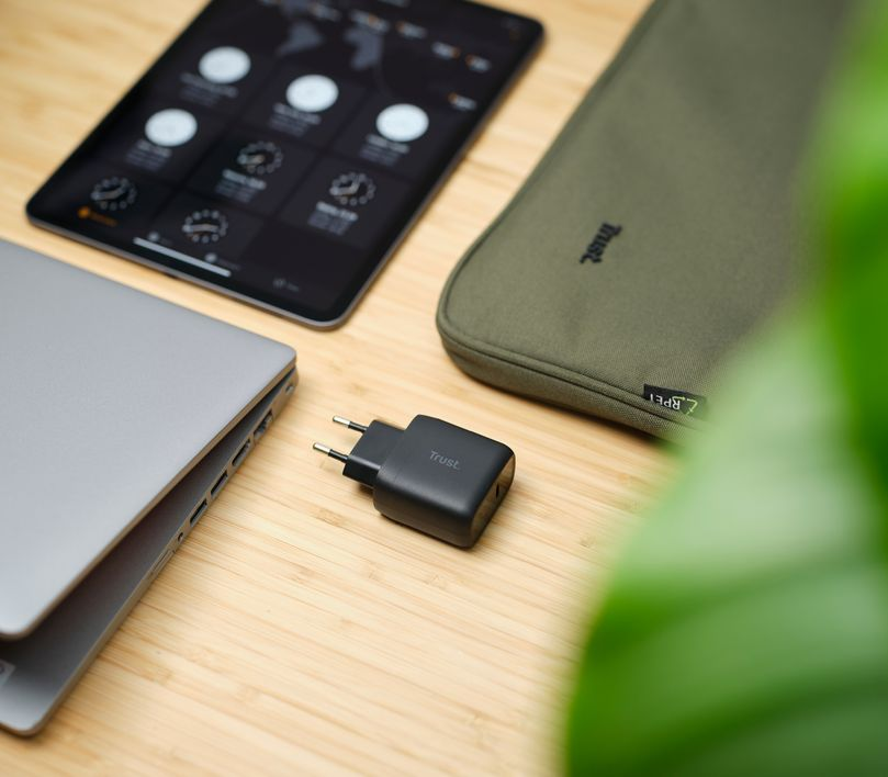 Egy laptop, tablet, töltő és egy zöld tok egy fa asztalon.