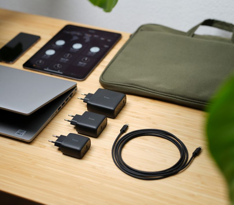 Fa asztal laptop, tablet, töltők és egy zöld tok. Egy fekete kábel van az asztalon.
