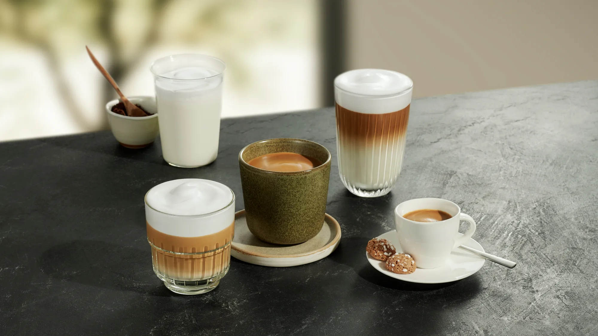 Verschiedene Kaffeespezialitäten in Tassen und Gläsern mit Keksen auf einem dunklen Tisch.