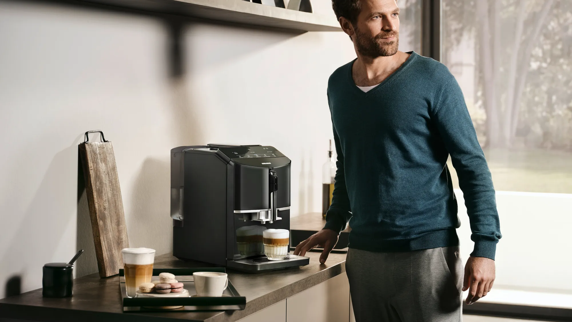 Ein Mann steht neben einer modernen Kaffeemaschine in einer Küche mit Kaffee und Macarons auf dem Tisch.