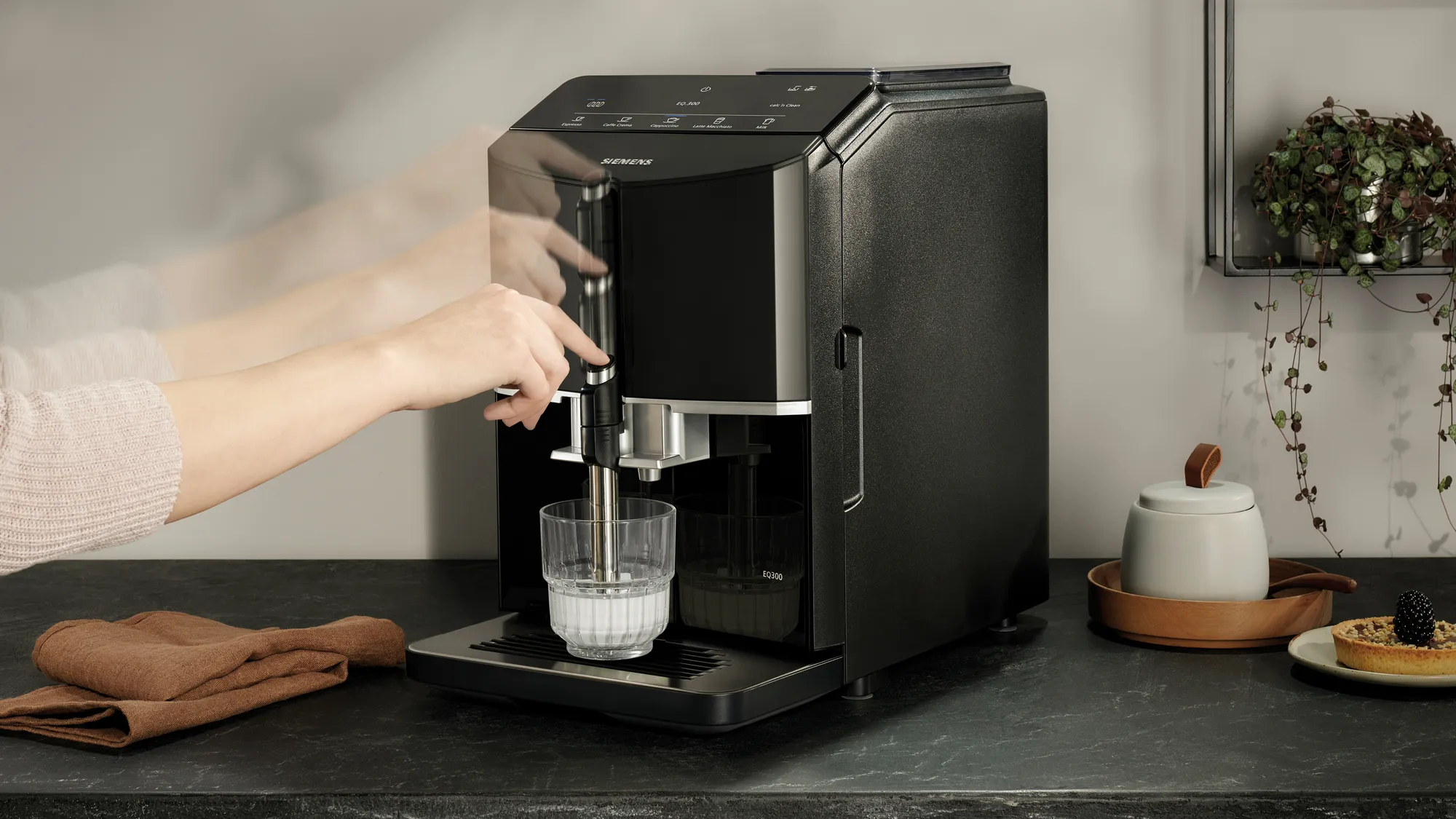 Eine Person bedient eine moderne schwarze Siemens-Kaffeemaschine auf einer dunklen Arbeitsplatte mit einem Glas Milch.