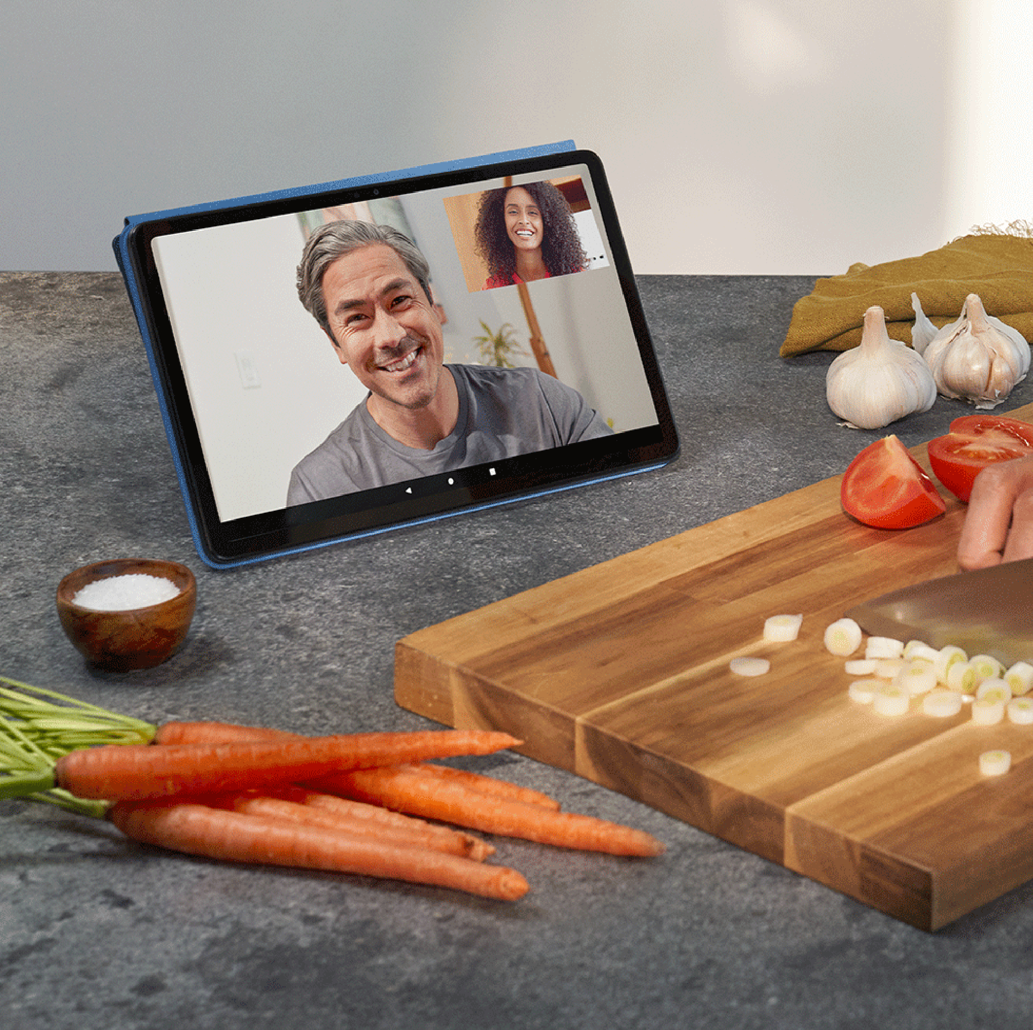 Tablet zeigt Videoanruf. Karotten, Tomaten, Knoblauch und Schneidebrett auf der Arbeitsplatte. Küchenszene.