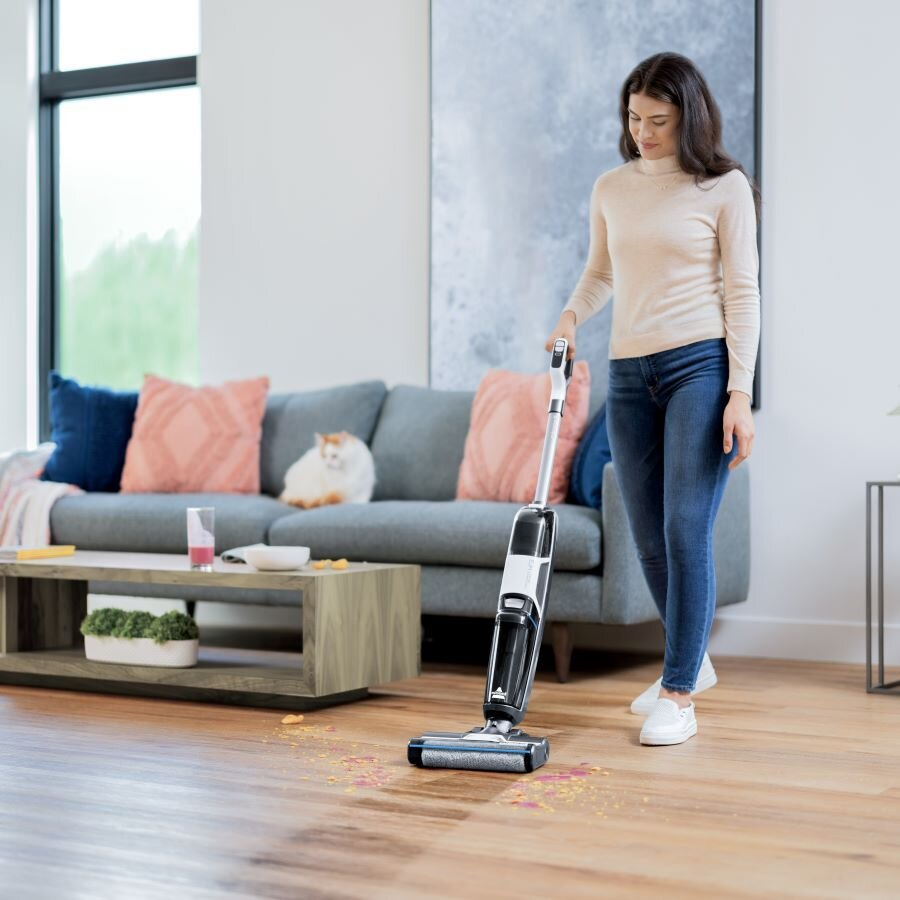 Kobieta odkurza drewnianą podłogę. Sofa, różowe poduszki, kot. Rozrzucone śmieci na podłodze.