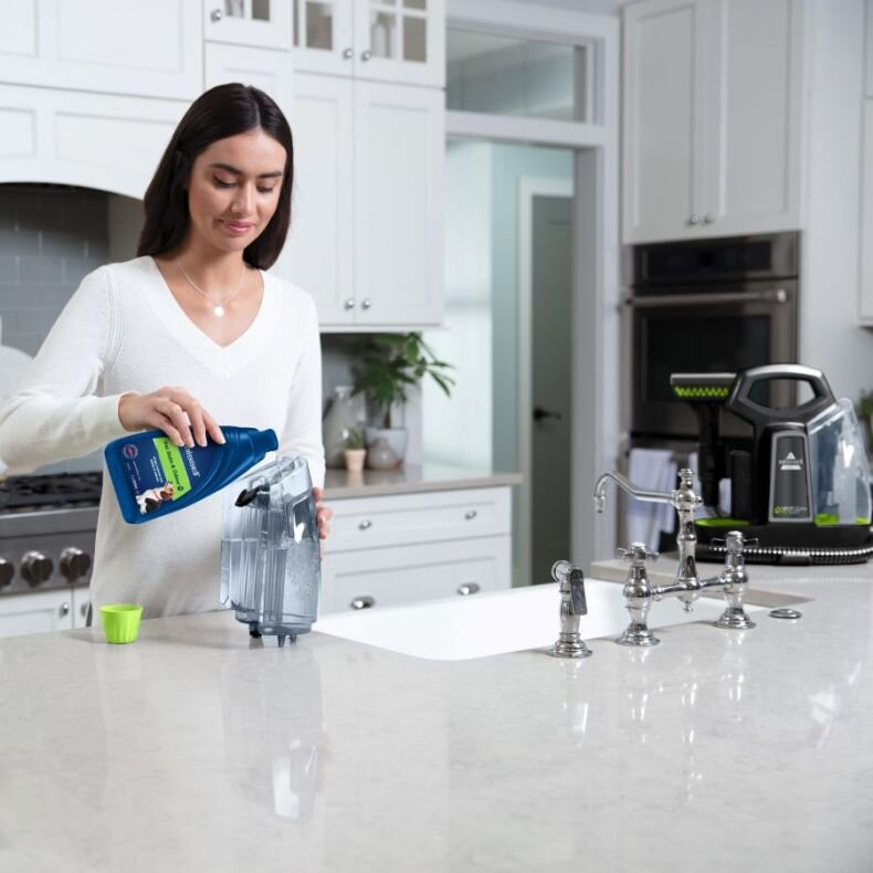 Kobieta wlewa roztwór czyszczący do zbiornika na wodę w białej kuchni. Widoczny jest środek czyszczący Bissell.