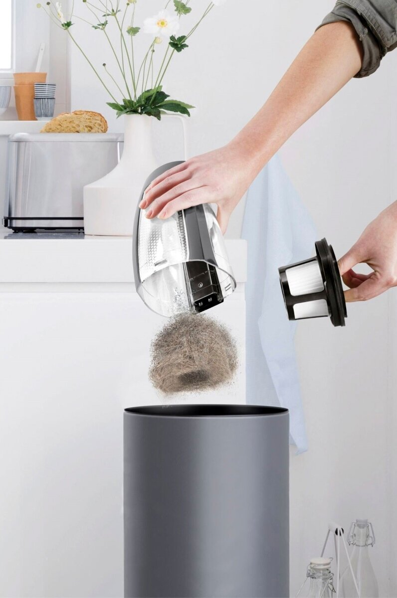 Ręce opróżniają pojemnik na kurz odkurzacza do szarego kosza. Widoczny kurz i filtr.