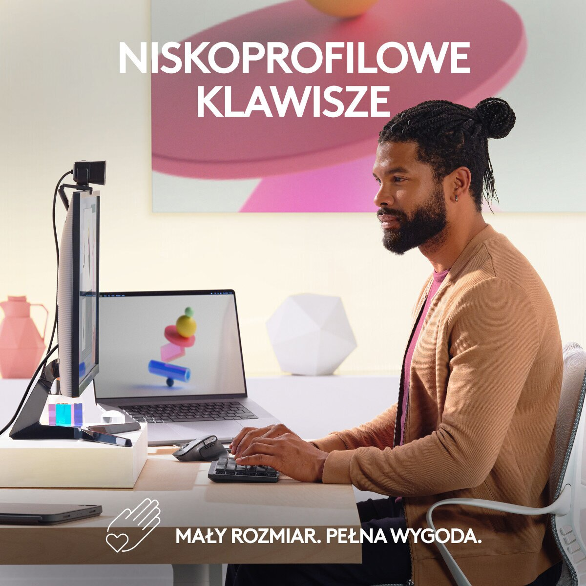 Mężczyzna z ciemnymi włosami siedzi przy biurku, pisząc na klawiaturze z laptopem i monitorem.