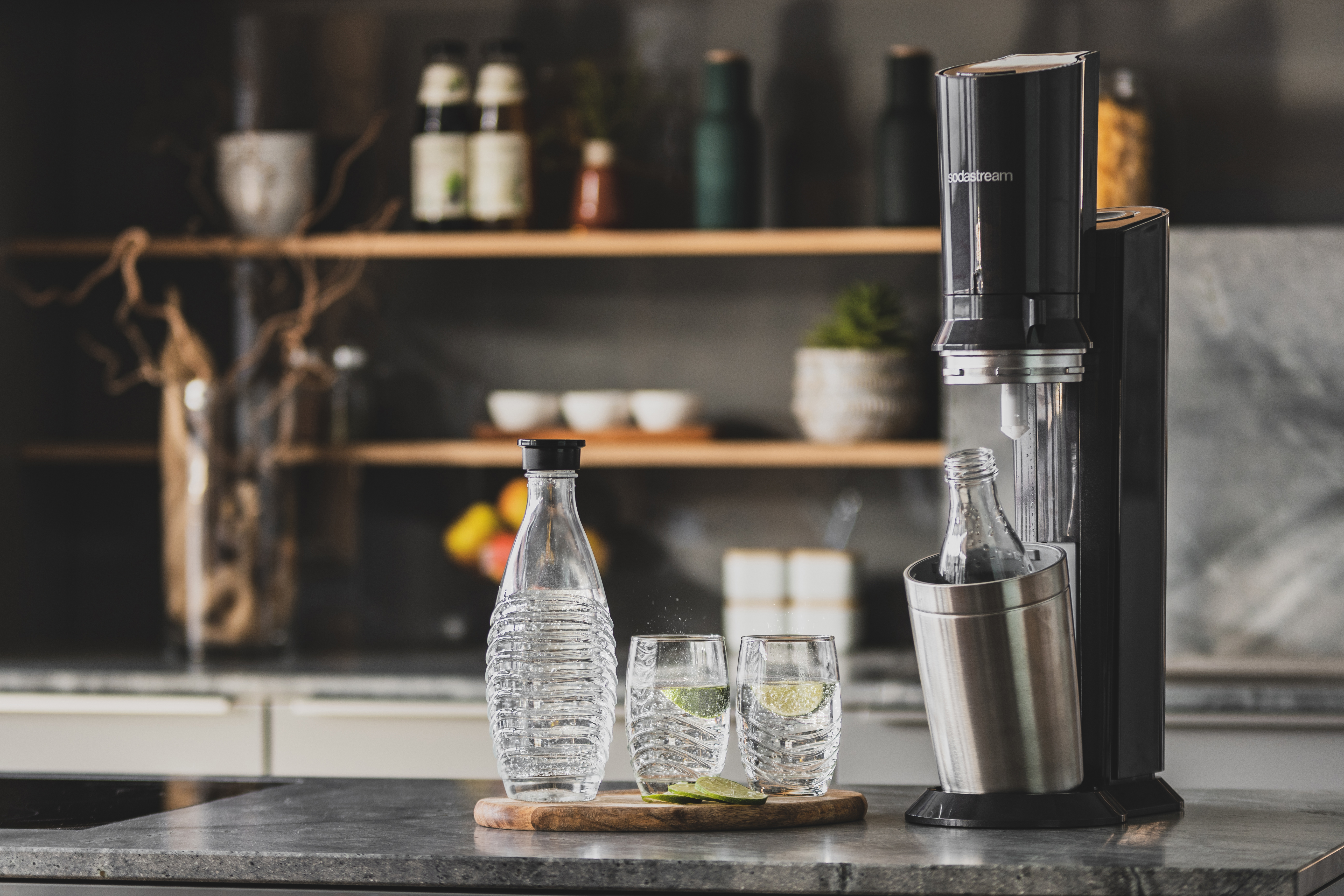 Une machine SodaStream avec une bouteille et deux verres avec des citrons verts et du soda sur un comptoir.