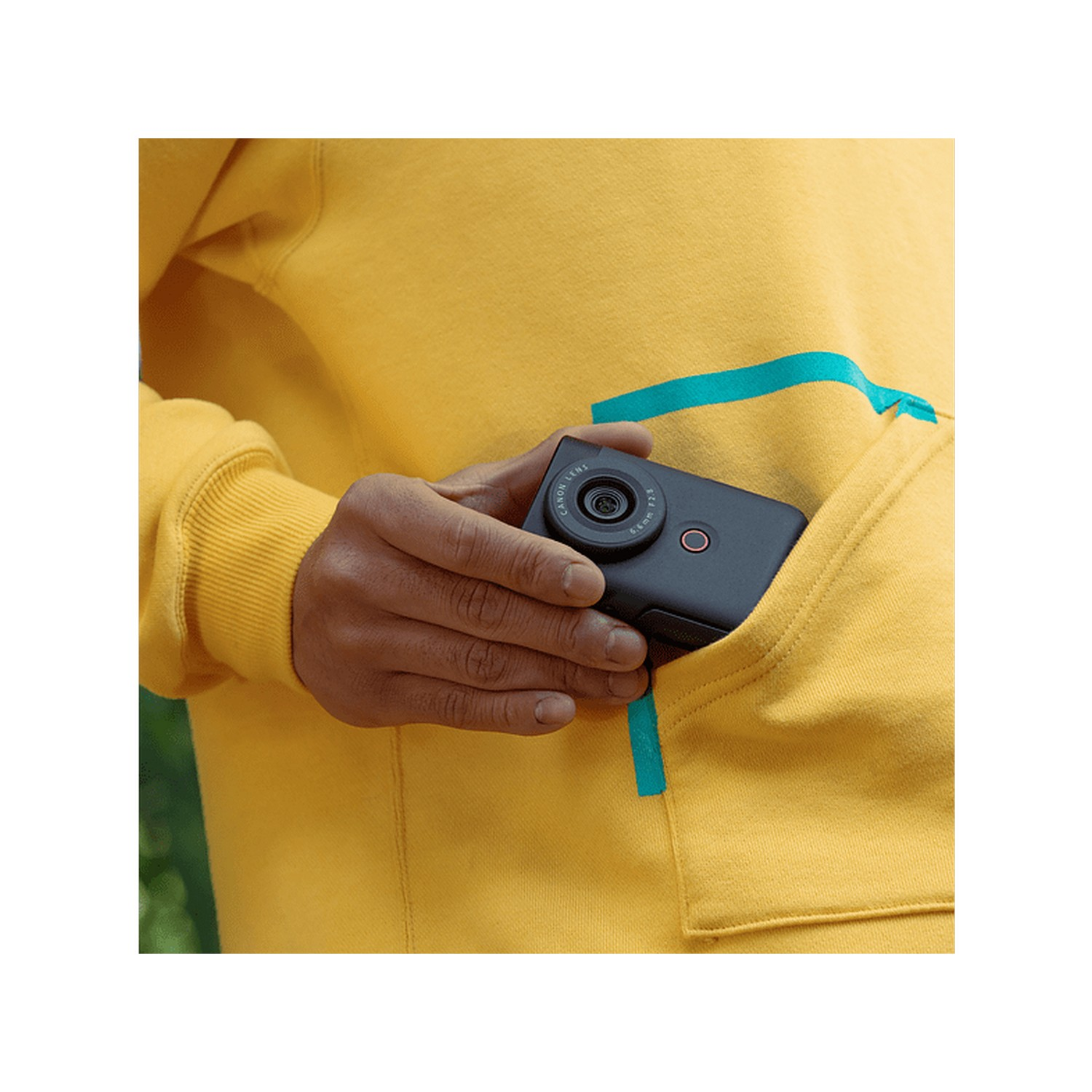 Person holding a black camera in a yellow hoodie pocket, showing the lens.