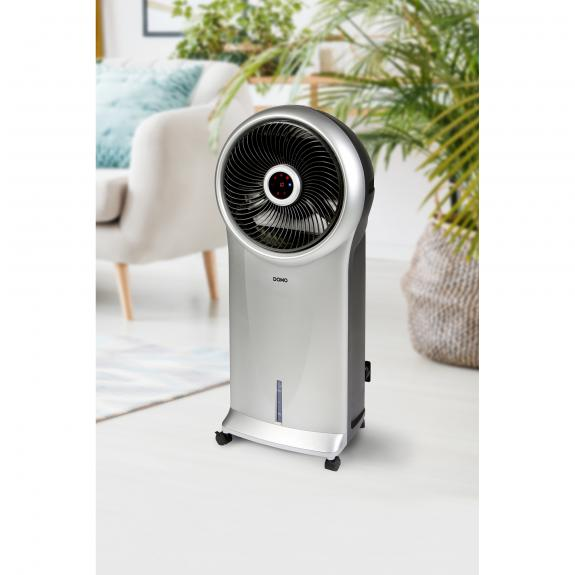 Un refroidisseur/ventilateur d'air argenté et noir sur un fond intérieur avec une chaise et des plantes.