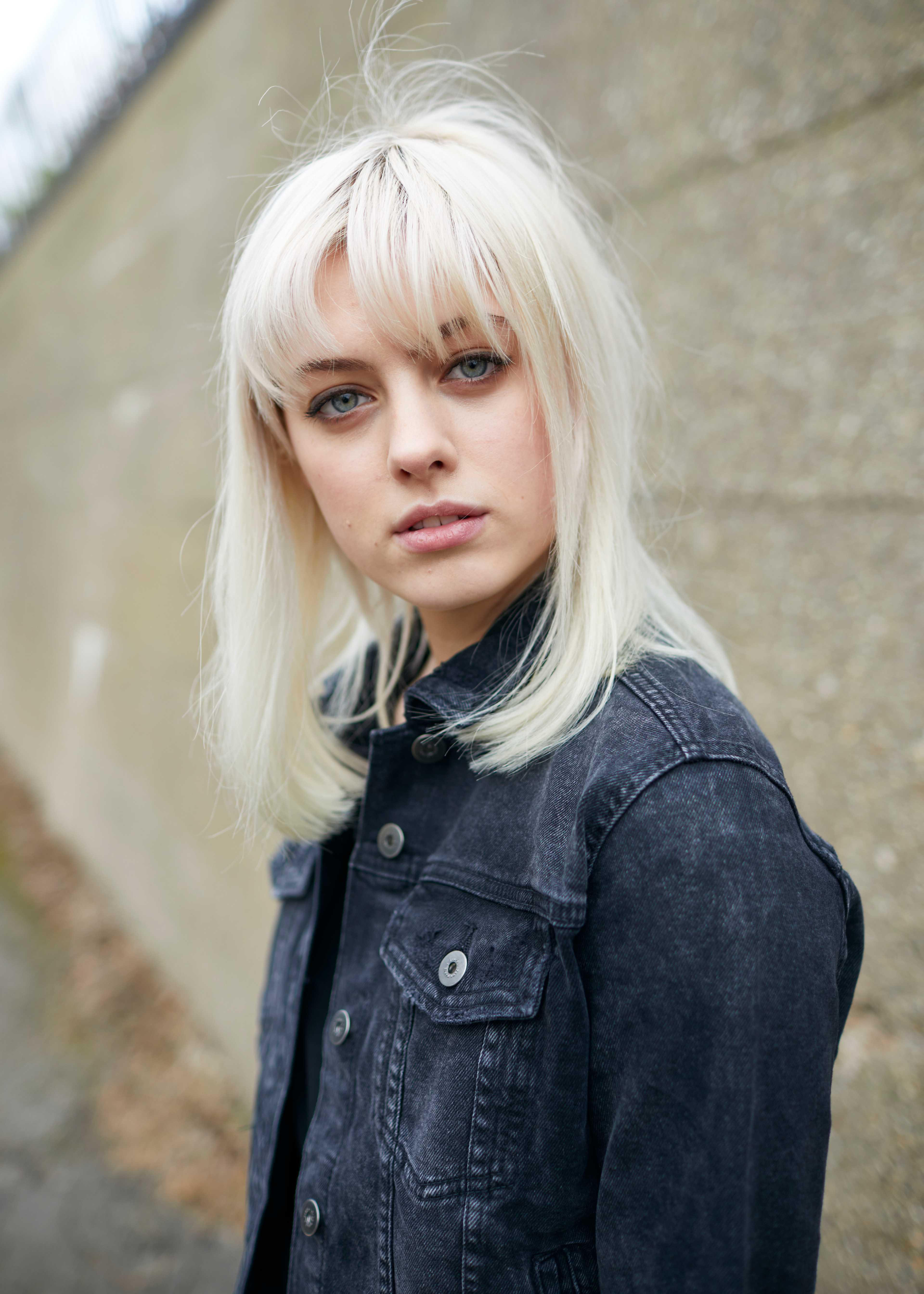 Eine junge Frau mit blonden Haaren in einer Jeansjacke blickt in die Kamera, im Freien.