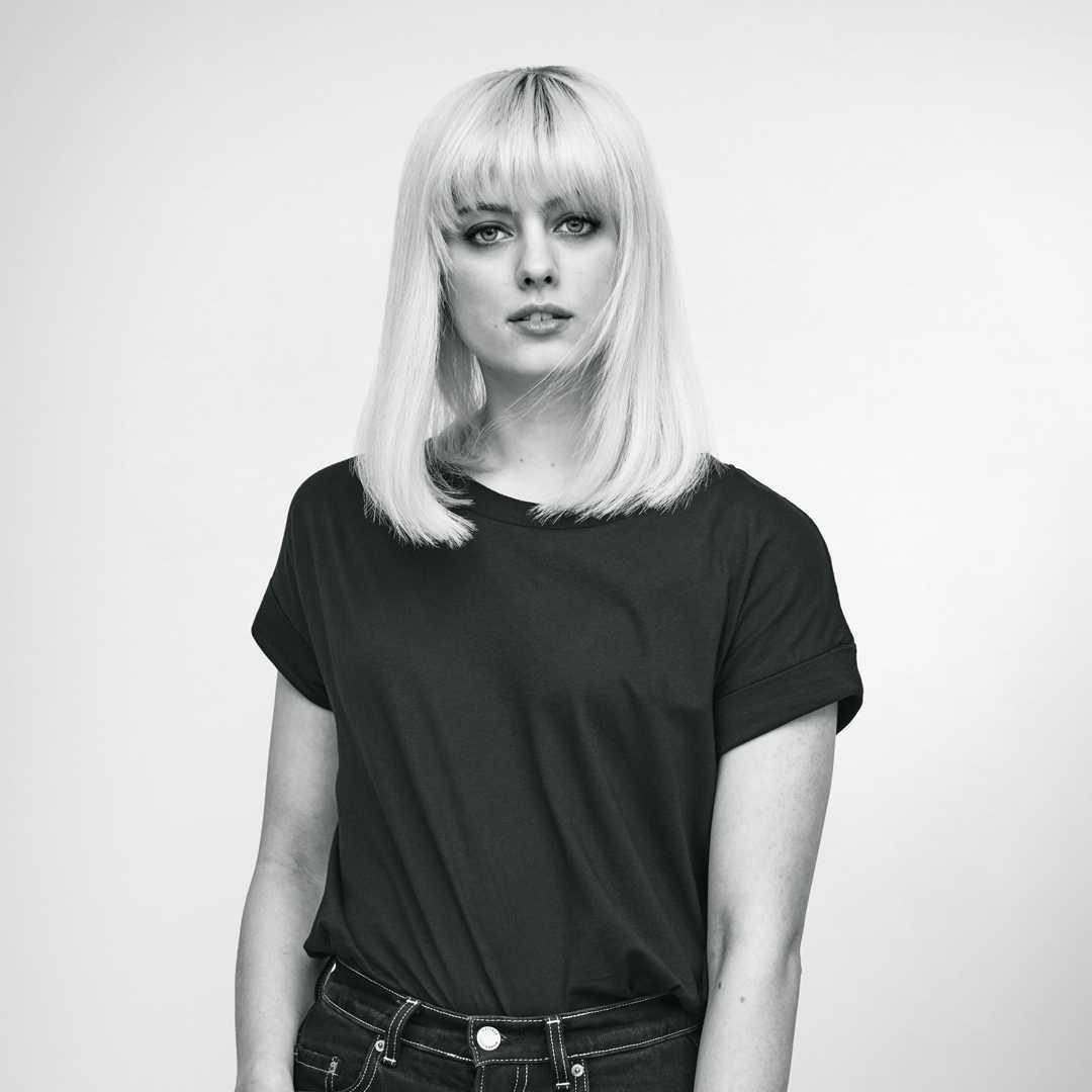 Eine Frau mit blonden Haaren und dunklem T-Shirt posiert vor weißem Hintergrund.