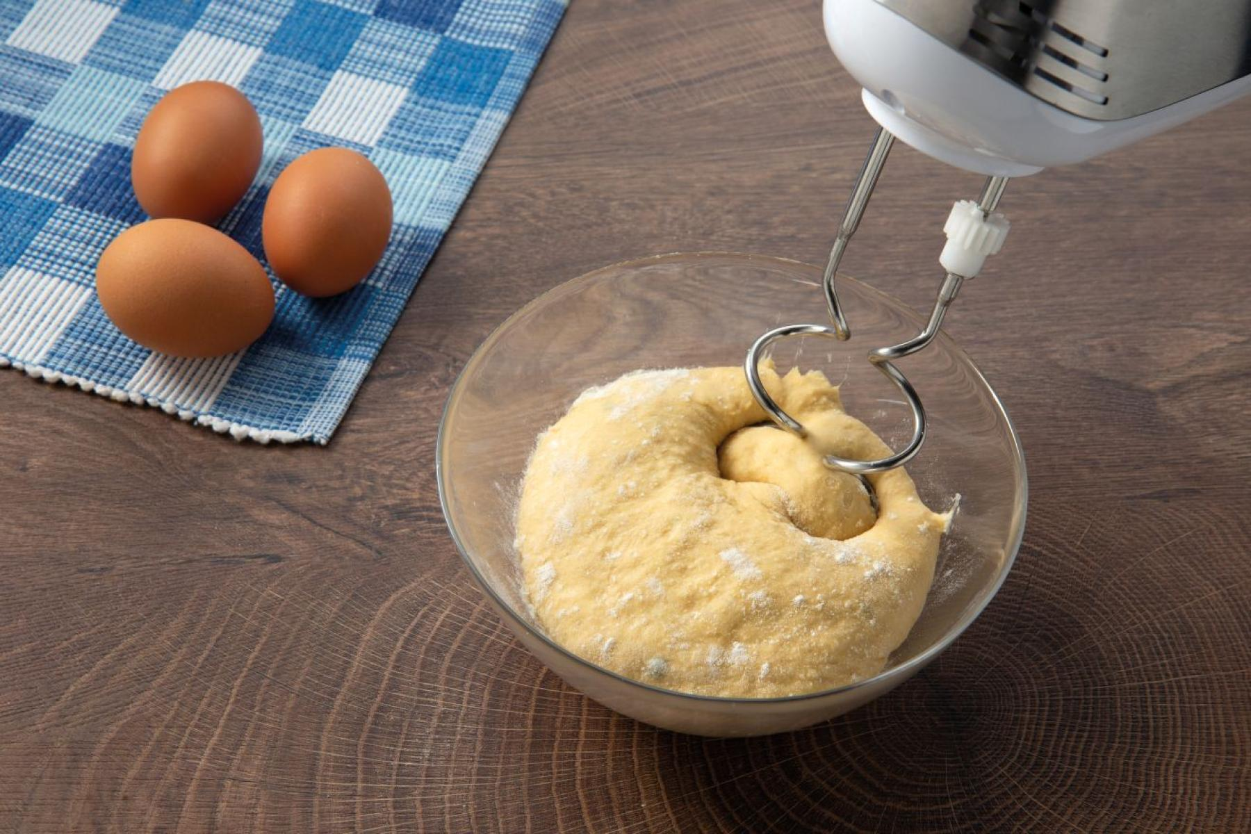 Tojások, tészta egy üvegtálban, kézi mixerrel keverve, és egy kék kockás kendő.