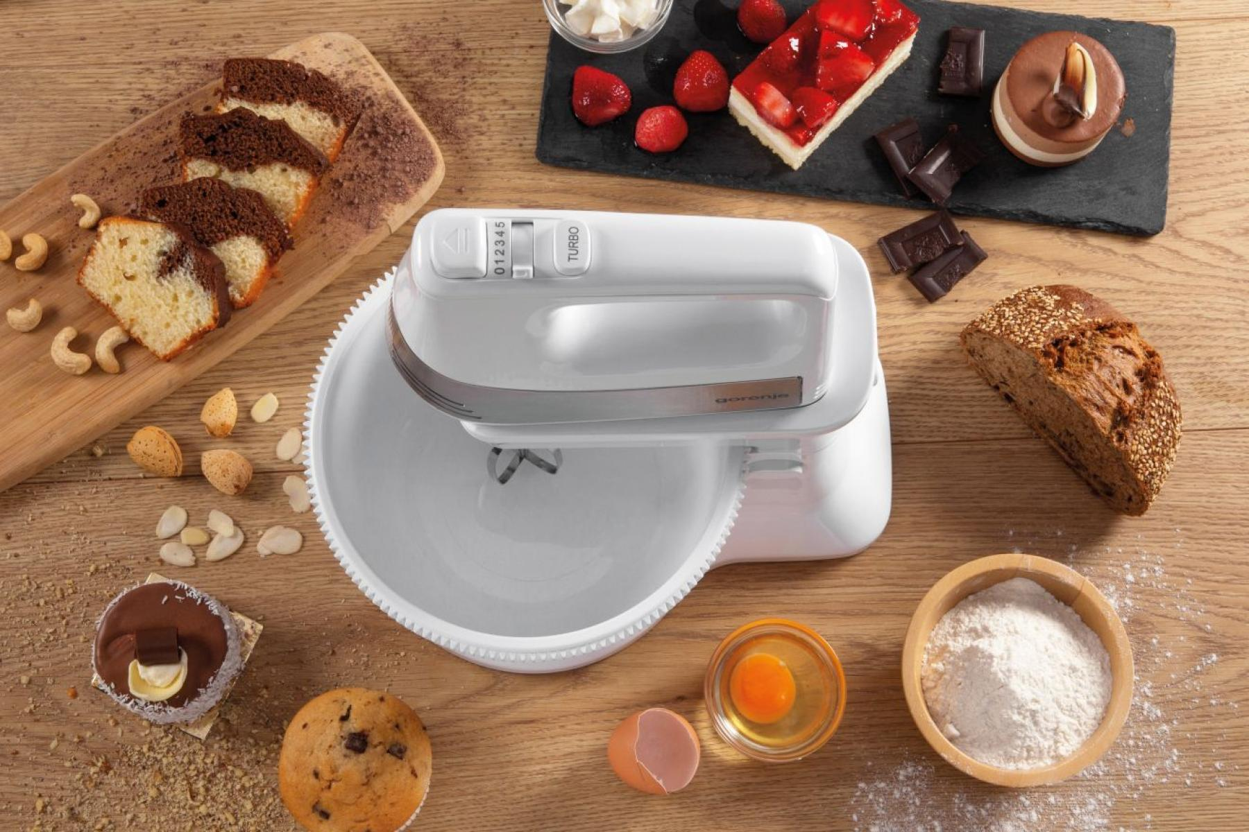 Egy fehér kézi mixer egy tálban, süteményekkel és hozzávalókkal körülvéve egy fa felületen.