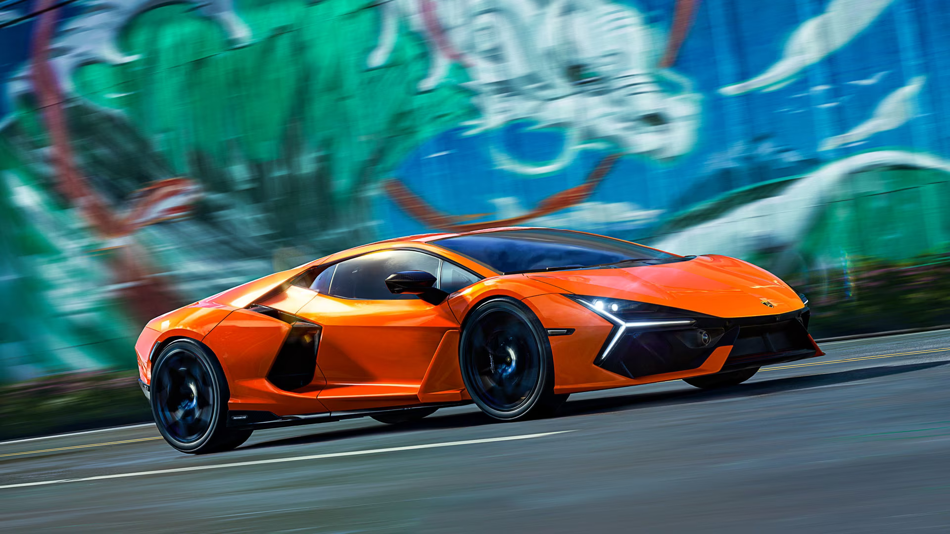 Un'auto sportiva arancione sfreccia su una strada, con un muro di graffiti sfocato sullo sfondo.