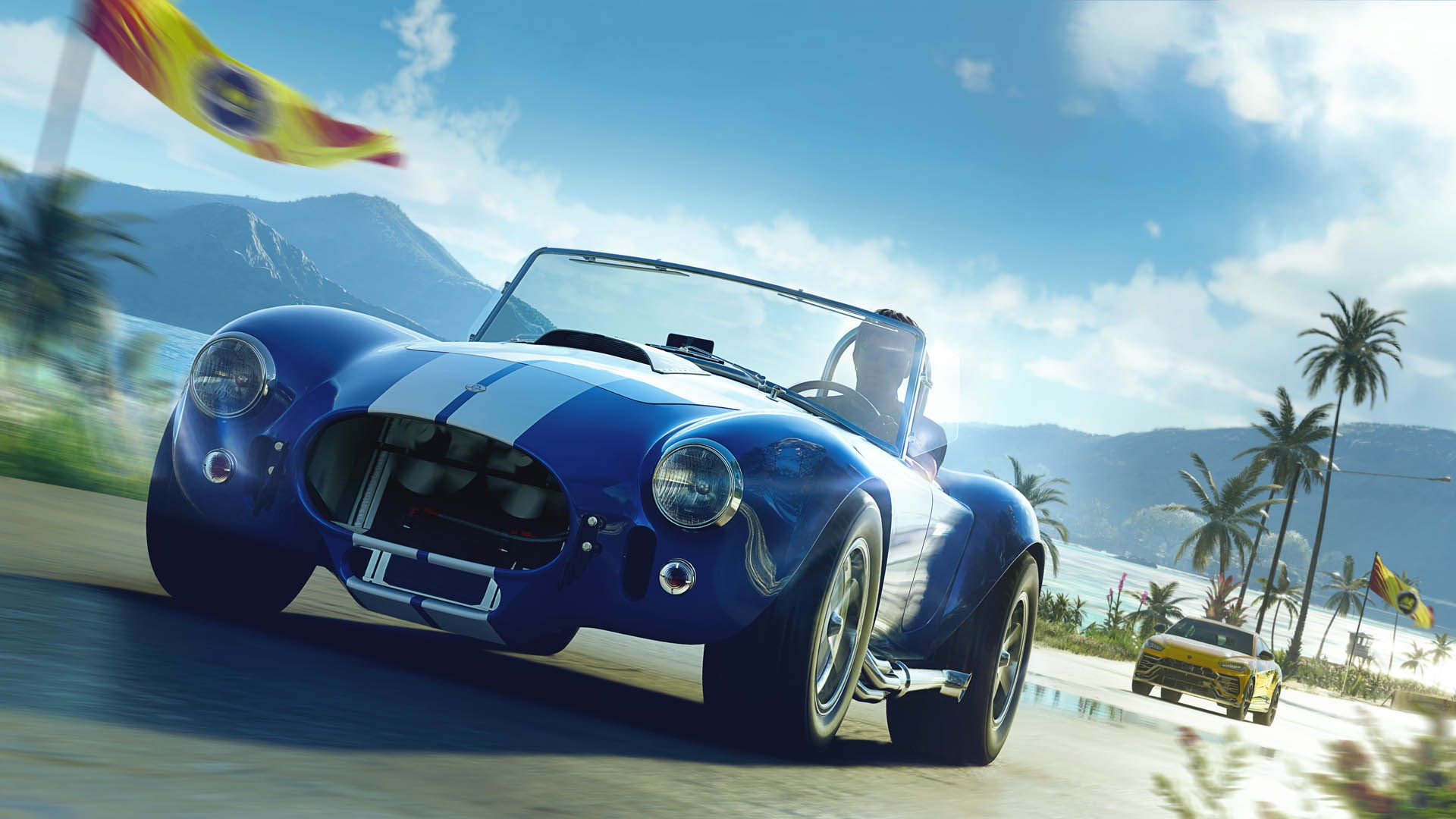 Un'auto sportiva Shelby Cobra blu e bianca sfreccia lungo una strada costiera con un'altra auto in lontananza.